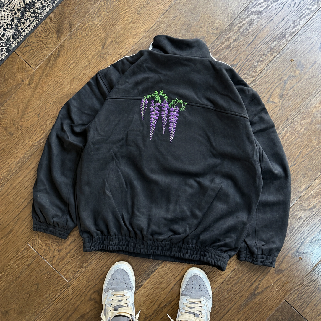 A black zip-up jacket with a purple wisteria flower graphic on the back, laid flat on a wooden floor. The bottom of a person's legs wearing gray and navy sneakers with white and yellow stripes is visible at the bottom of the frame.