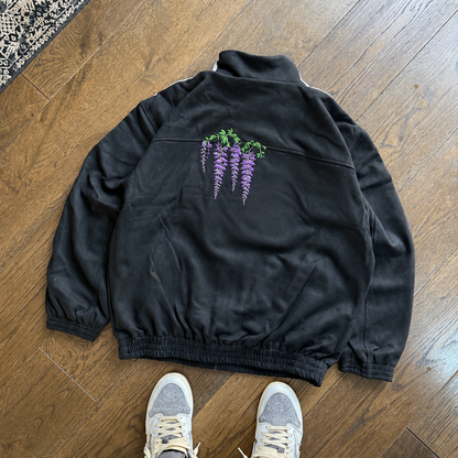 A black zip-up jacket with a purple wisteria flower graphic on the back, laid flat on a wooden floor. The bottom of a person's legs wearing gray and navy sneakers with white and yellow stripes is visible at the bottom of the frame.