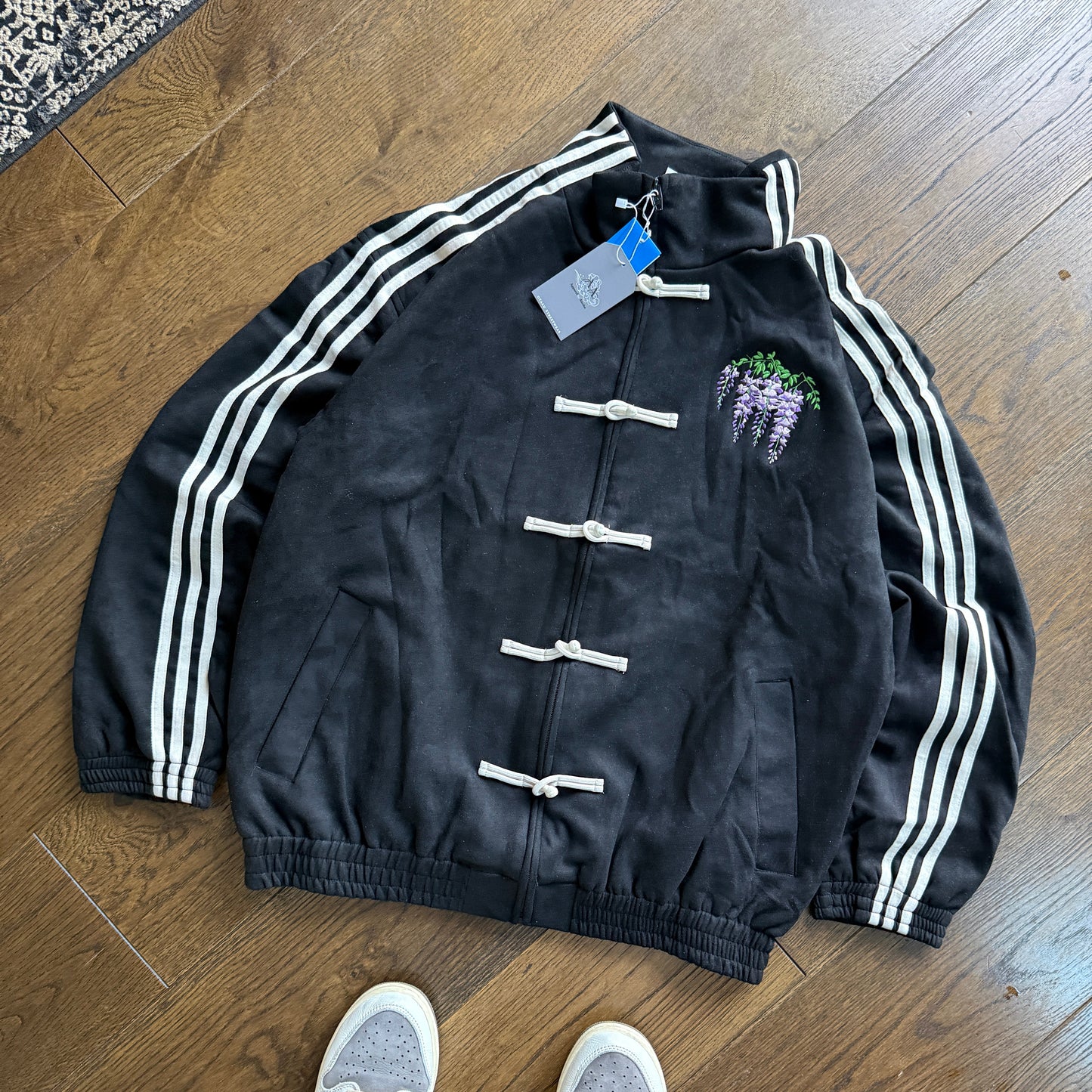 A black track jacket with white stripes down the sleeves and white toggle closures, laid flat on a wooden floor. A person's feet wearing gray sneakers are visible at the bottom of the frame.