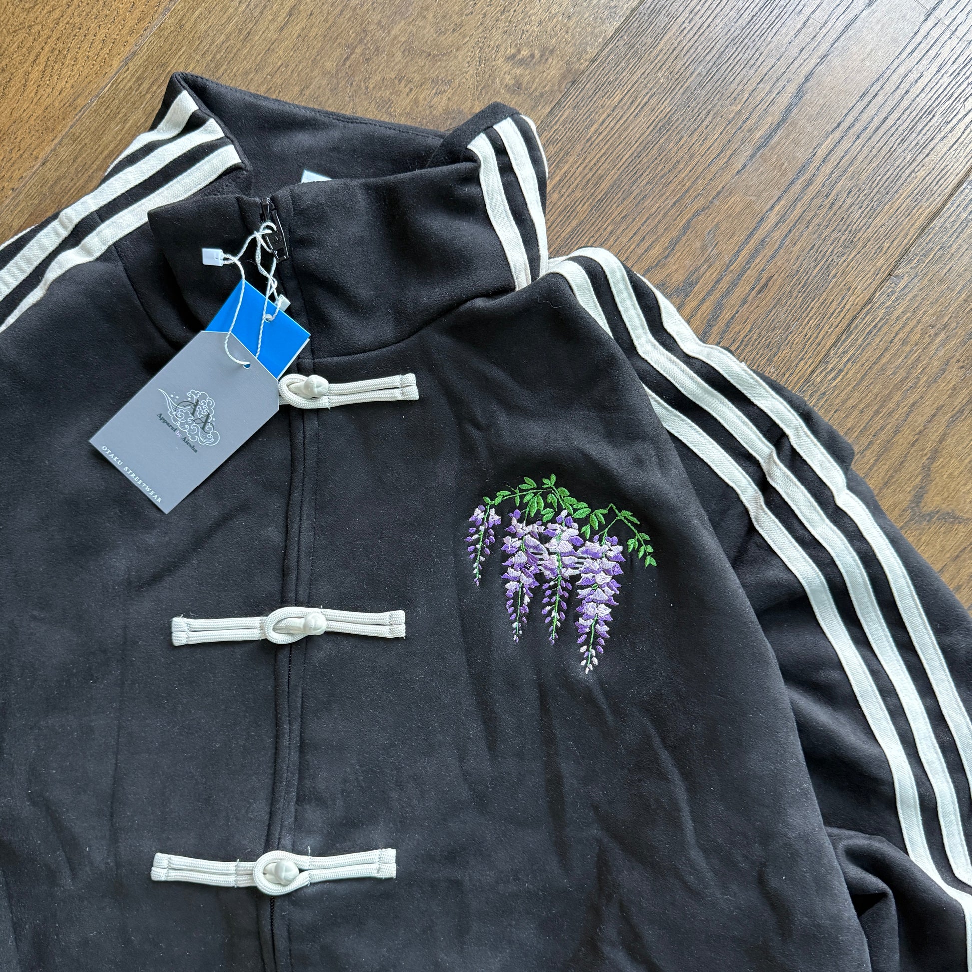 A black velour track jacket with white three-stripes on the sleeves, featuring white Chinese-style toggle closures and an embroidered purple wisteria flower on the chest. A gray brand tag is attached to the collar, and the jacket is laid flat on a wooden floor.