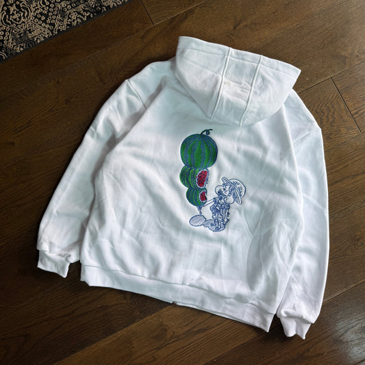 A white long-sleeve sweatshirt with a graphic of watermelons and a blue cat, laid flat on a dark wood floor.