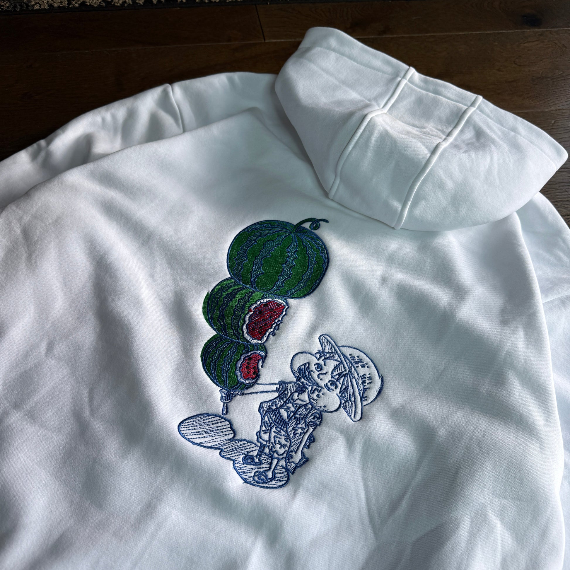 A white long-sleeve hoodie laid flat on a dark wood floor, featuring a graphic print of green watermelons with red flesh and a blue-and-white cartoon character.