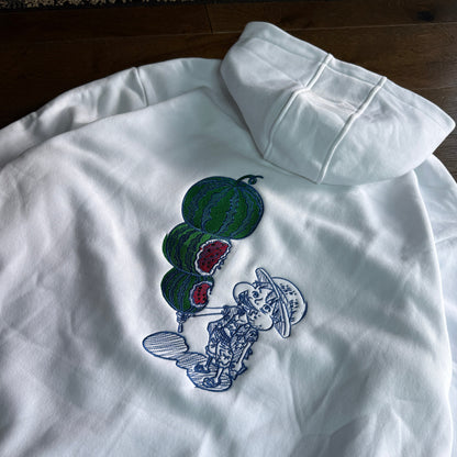 A white long-sleeve hoodie laid flat on a dark wood floor, featuring a graphic print of green watermelons with red flesh and a blue-and-white cartoon character.