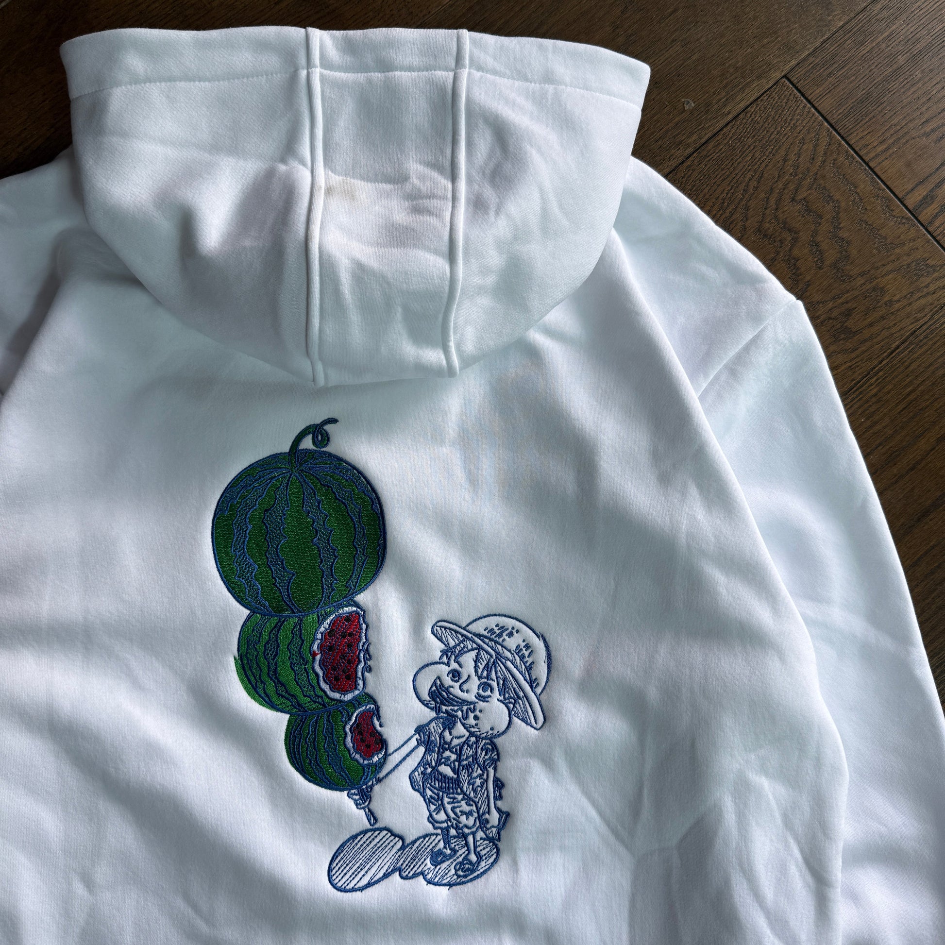 A white hooded sweatshirt laid flat on a dark wood floor, featuring a large embroidered graphic on the back of a cartoon character holding watermelons.