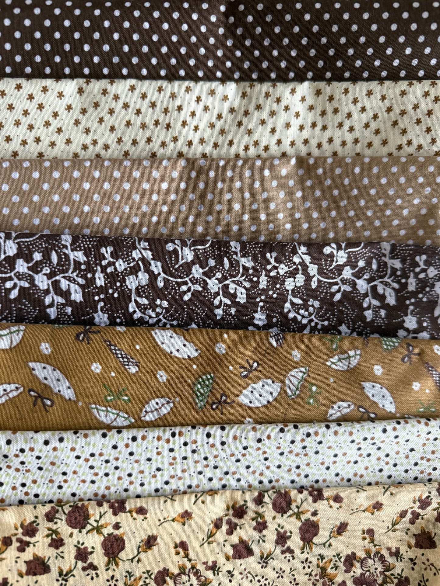 A stack of folded fabric samples with various patterns, including polka dots, floral prints, and leaf motifs in earthy brown, beige, and cream tones.
