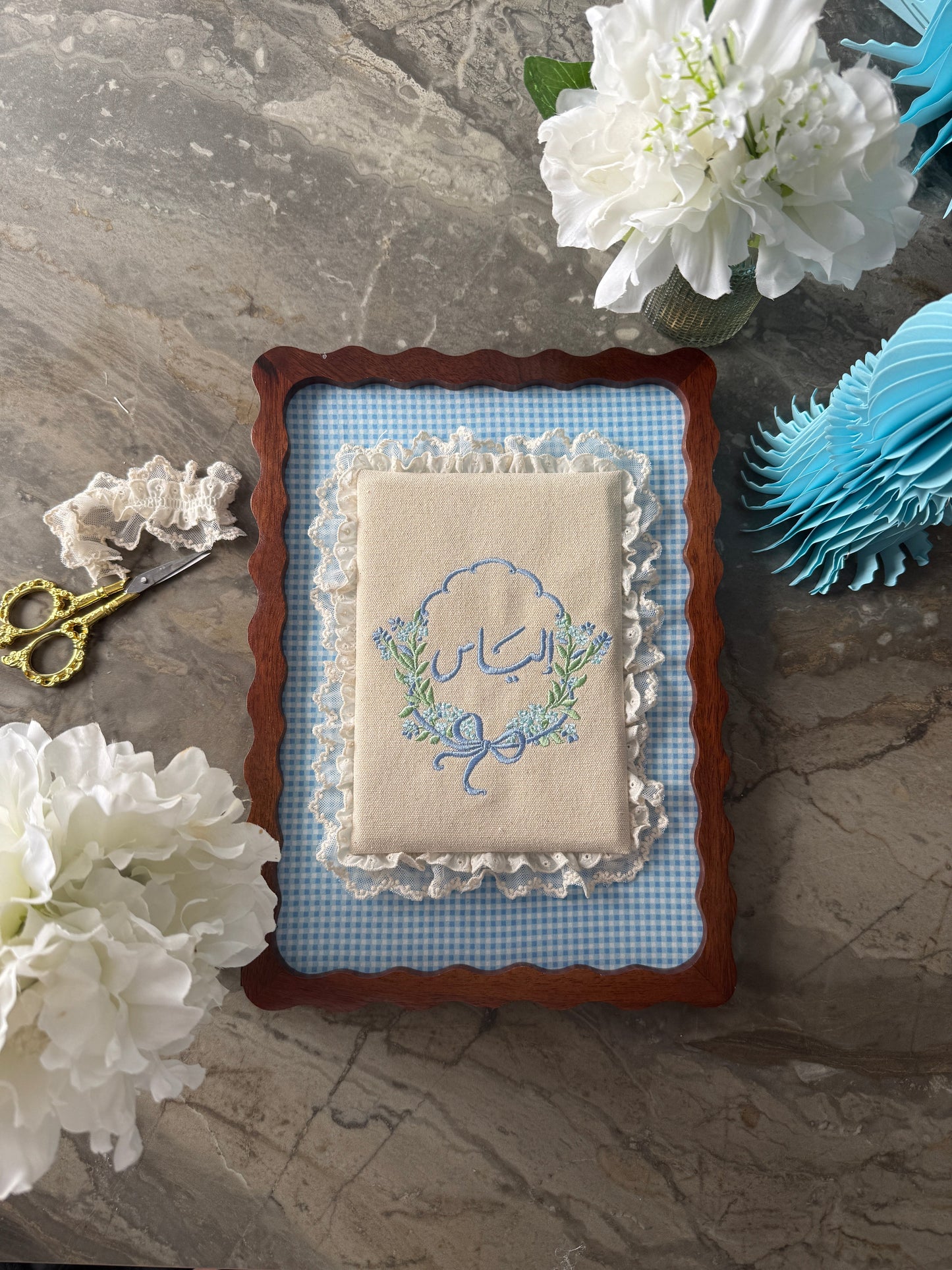 An embroidered baby keepsake pillow with a blue and green floral design, set in a scalloped brown wooden frame with a blue gingham mat. The pillow is styled on a gray marble surface with white flowers, gold scissors, and a blue paper fan.