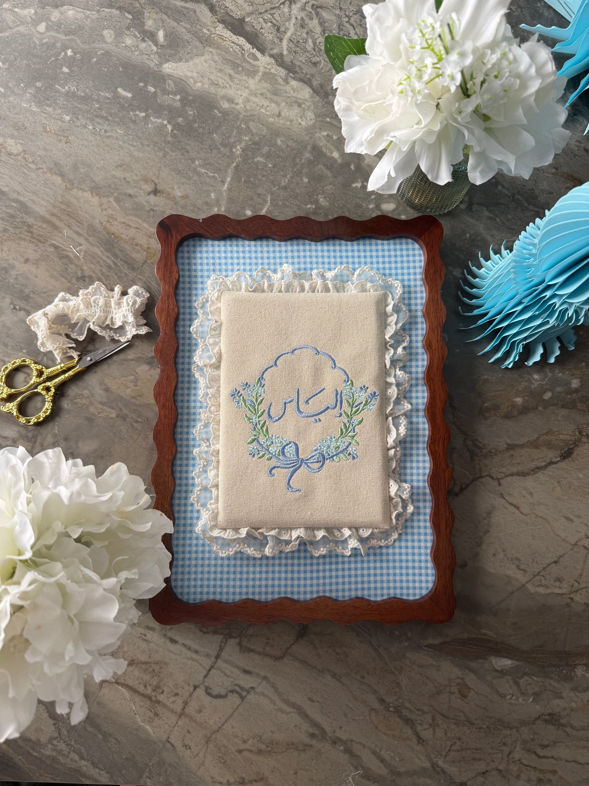 An embroidered baby keepsake pillow with a blue and green floral design, set in a scalloped brown wooden frame with a blue gingham mat. The pillow is styled on a gray marble surface with white flowers, gold scissors, and a blue paper fan.